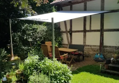 Helles Sonnensegel über einem Gartentisch mit Stühlen vor einer Fachwerkwand, ringsum Beete und Rasen