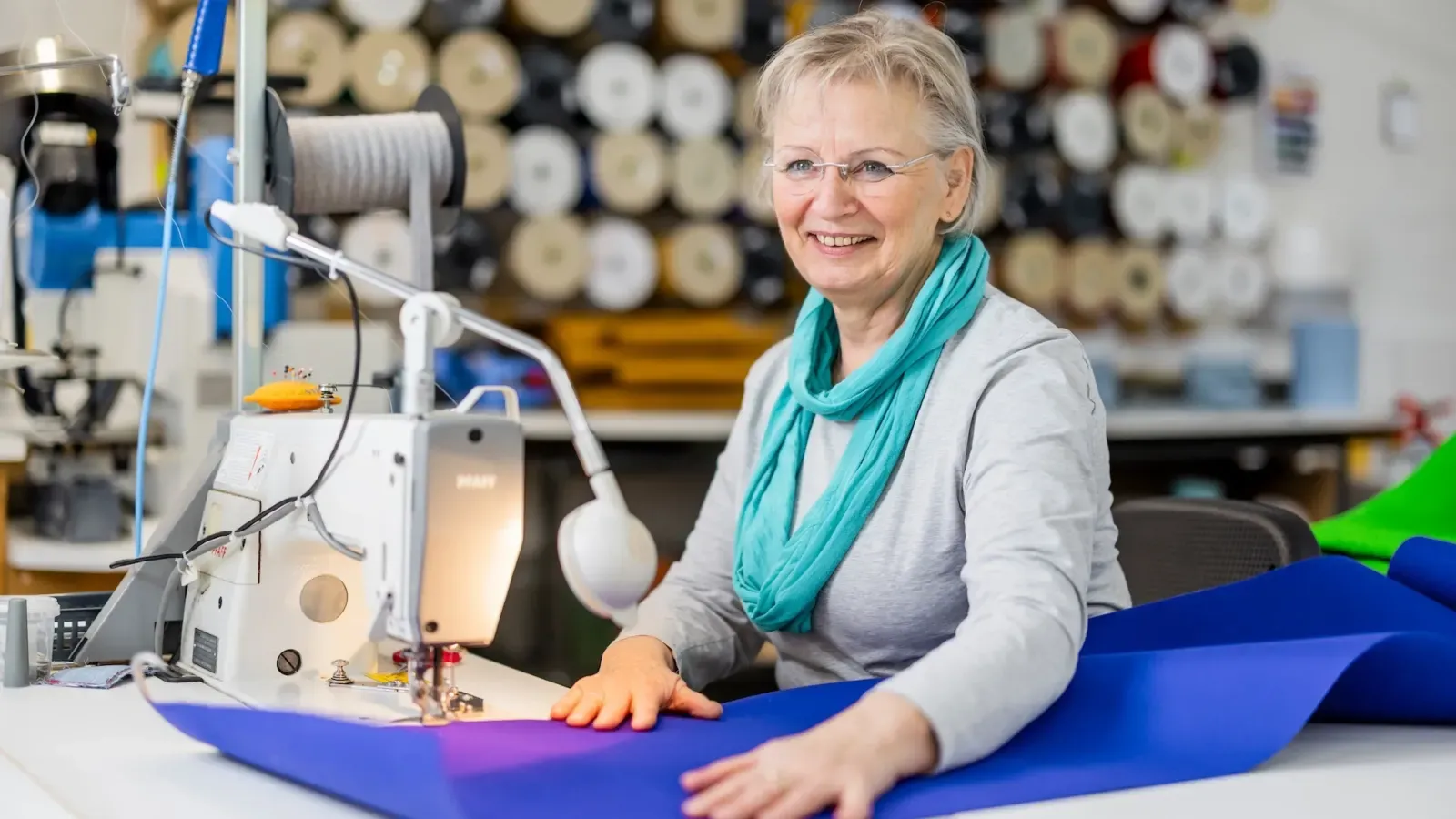 Mitarbeiterin führt blauen Markisenstoff an einer Industrienähmaschine, dahinter eine Wand mit Garnrollen