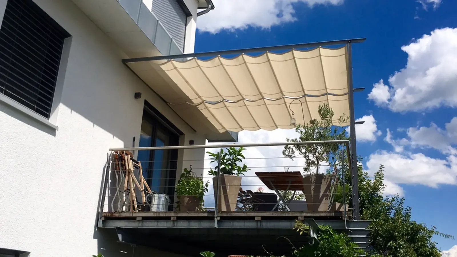 Beige Seilspannmarkise spannt sich über einen Balkon mit Tisch, Stühlen und Kübelpflanzen
