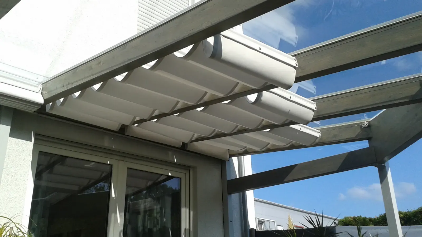 Weiße Stoffbahnen einer Seilspannmarkise hängen gerafft unter einem verglasten Terrassendach