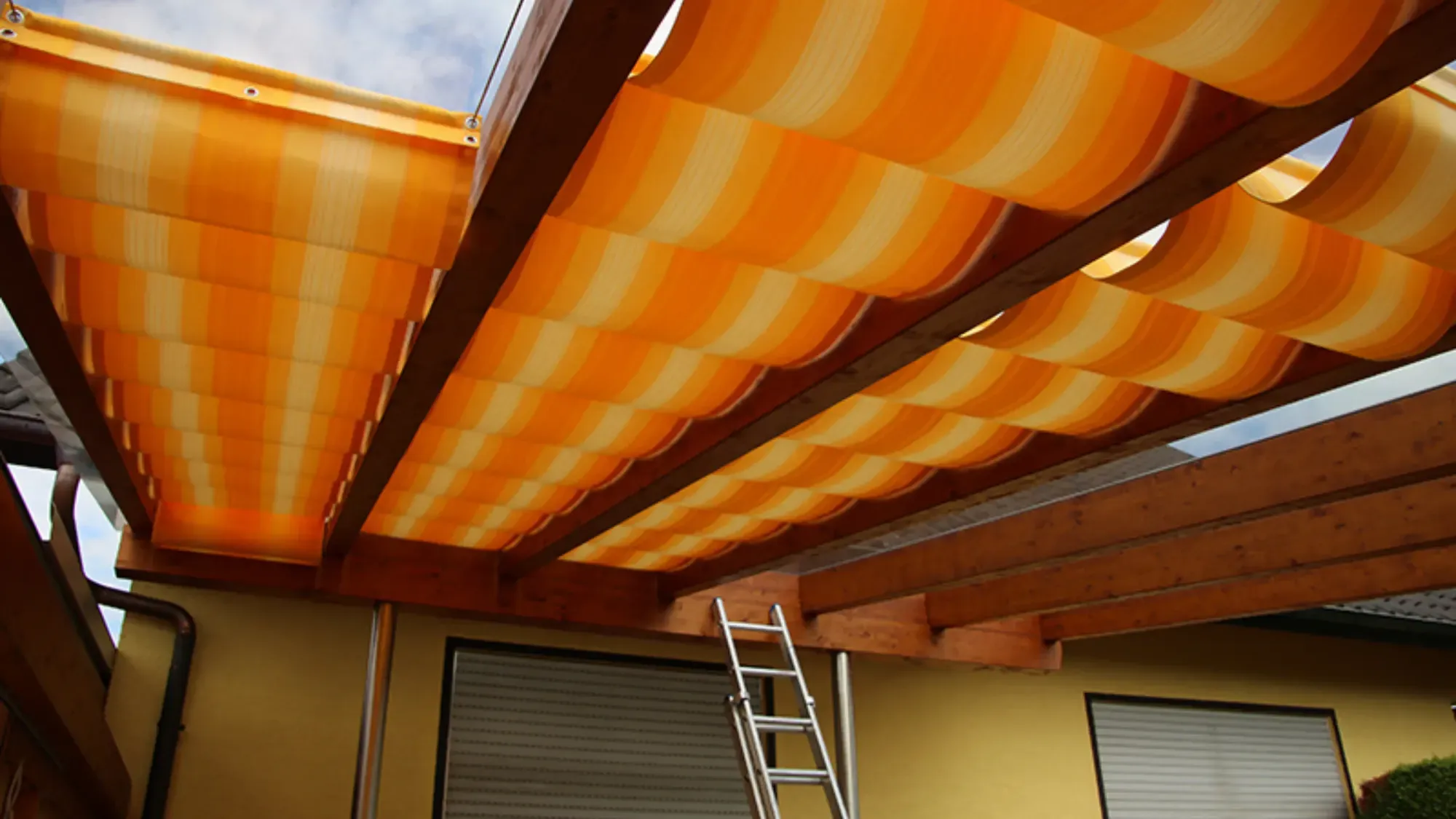 Blick von unten auf gelb-orange gestreifte Stoffbahnen zwischen den Holzsparren einer Pergola, rechts lehnt eine Aluleiter
