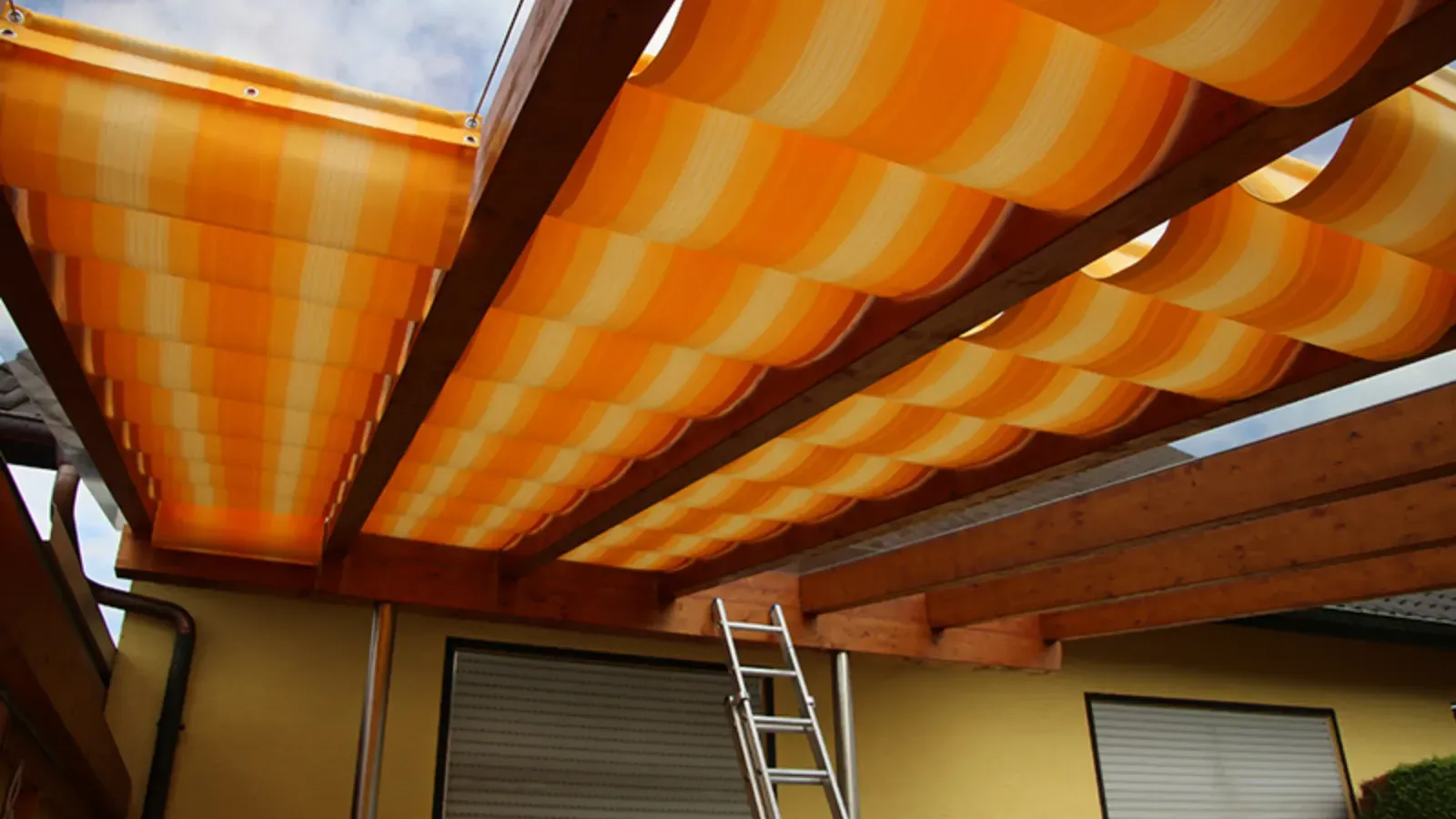 Gelb-orange gestreifte Stoffbahnen laufen unter den Holzsparren einer Pergola an der Hauswand