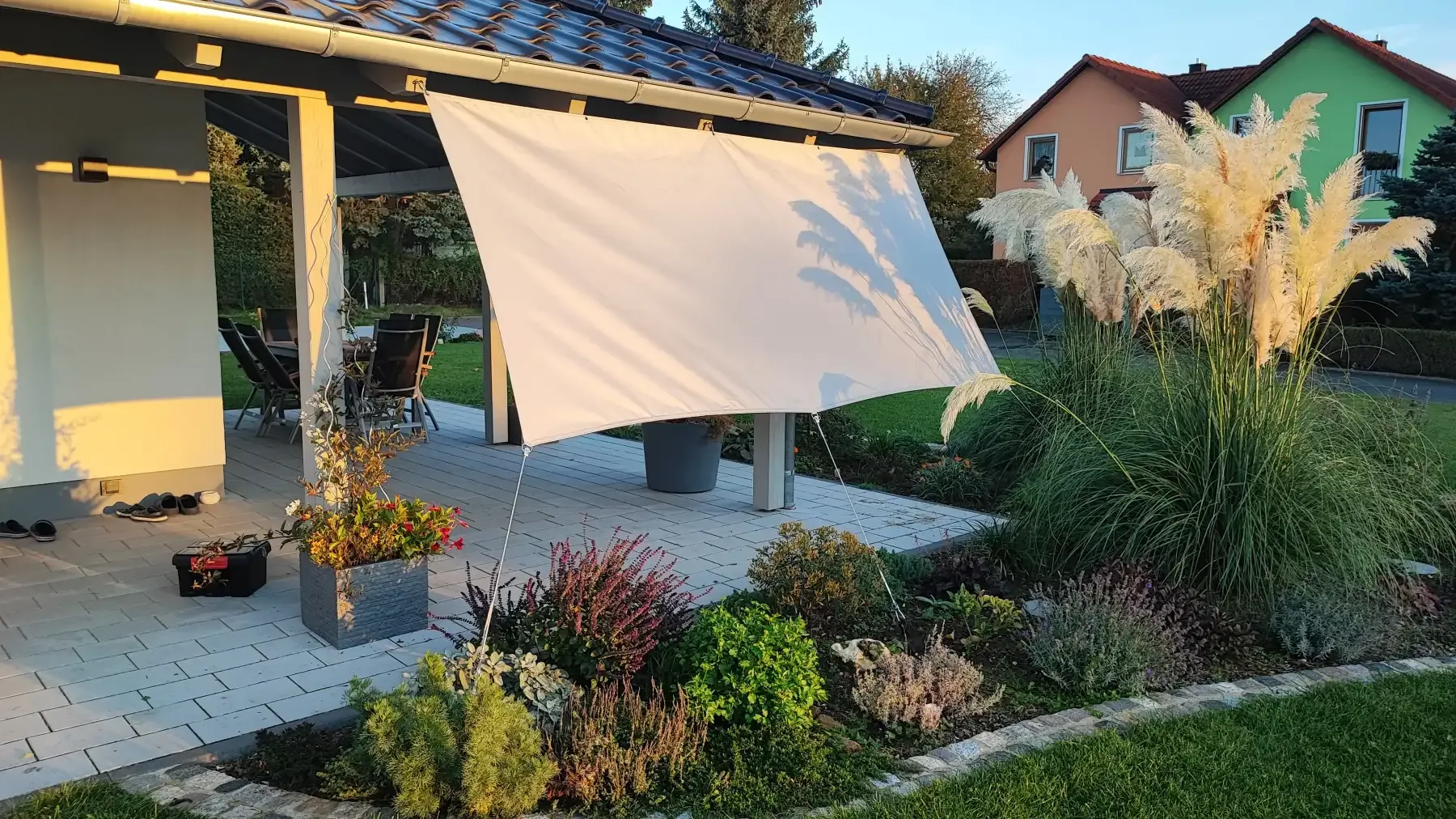 Senkrecht gespanntes helles Tuch zwischen Terrassendach und Bodenpunkten schirmt eine Sitzecke gegen tiefe Sonne ab